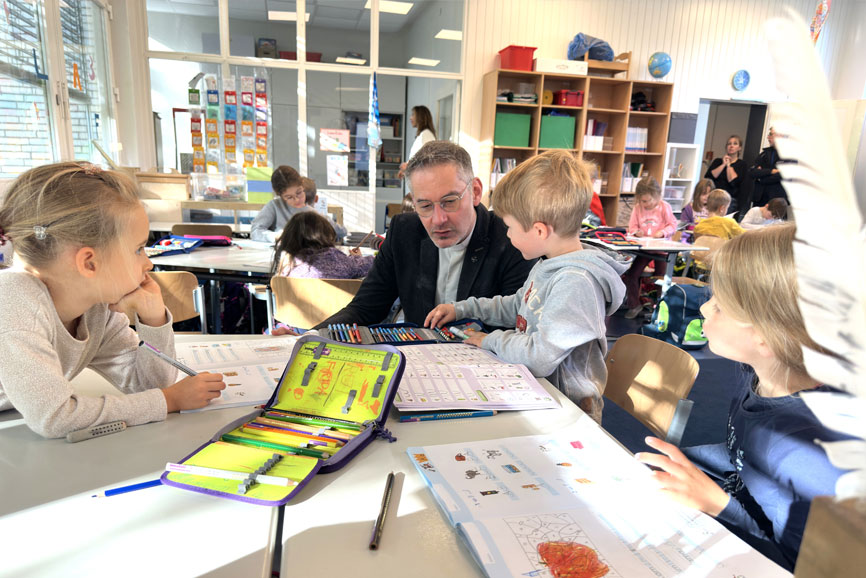 Generalvikar Geißler ist zu Besuch an der Katharina-von-Siena Schule in Langenhorn. Er sitz an einem Tisch in einer Klasse und um ihn rum sitzen ein paar Schüler im Alter von zirka 6 Jahren und sie unterhalten sich.