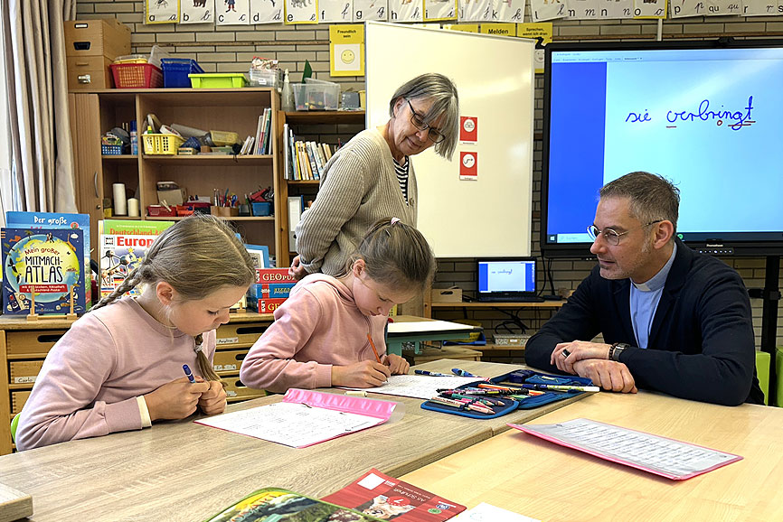 Generalvikar Geißler ist zu Besuch an der Katholischen Schule St. Joseph in Wandsbek und sitzt mit zwei Mädchen am Schreibtisch. Die beiden Mädchen schreiben etwas in ihre Schulhefte.