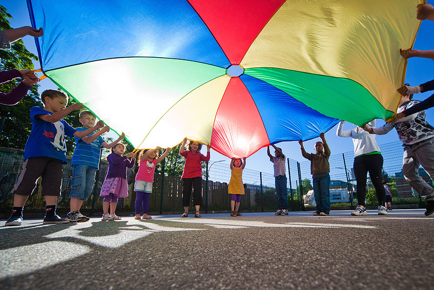 Das Bild zeigt viele Grundschulkinder mit einem regenbogenfarbenen, großen Tuch. Dieses schwingen die Kinder hoch und runter. Es ist strahlend blauer Himmel und die Sonne scheint.