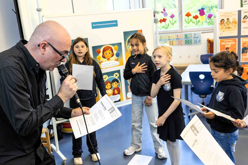 Ein kahlköpfiger Mann singt in ein mikrophon, drei Kinder stehen im Halbkreis um ihn herum und singen mit