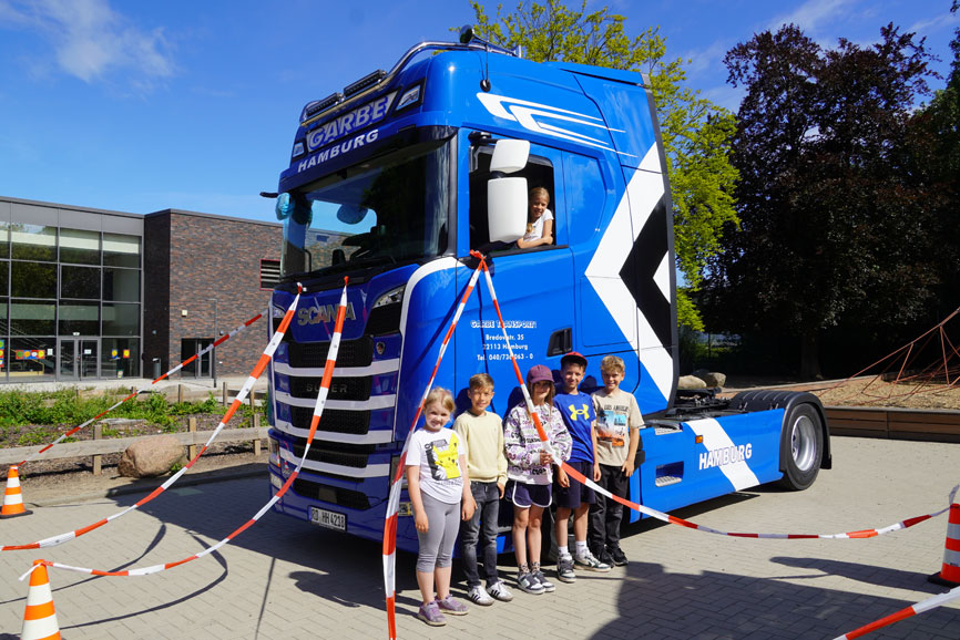 Ein 40-Tonner auf dem Schulhof - An der Katholischen Schule Farmsen war das kein Verkehrsverstoß, sondern praxisnaher Unterricht mit Weitblick - drei vierten Klassen der Grundschule am Rahlstedter Weg nahmen jetzt an einem besonderen Verkehrssicherheitstraining teil und hier zu sehen ist ein großer blauer LKW. Davor stehen 5 SchülerInnen und lehnen sich an den 40-Tonner.