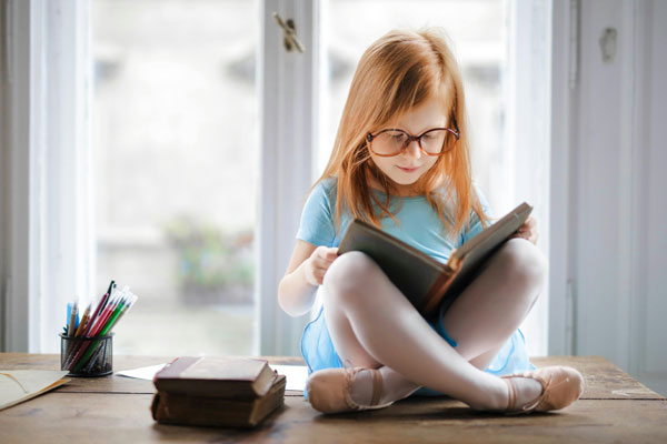 Ein kleines Mädchen sitzt im Schneidersitz auf einem Schreibtisch und schaut sich mit einer großen Brille auf der Nase ein Buch an.