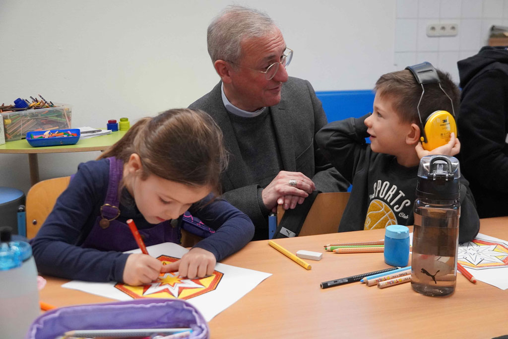 Erzbischof Heße zu Gast an der Katholischen Schule Hammer Kirche - hier zu sehen im Klassenraum einer Grundschulklasse. Er unterhält sich mit einem Jungen.