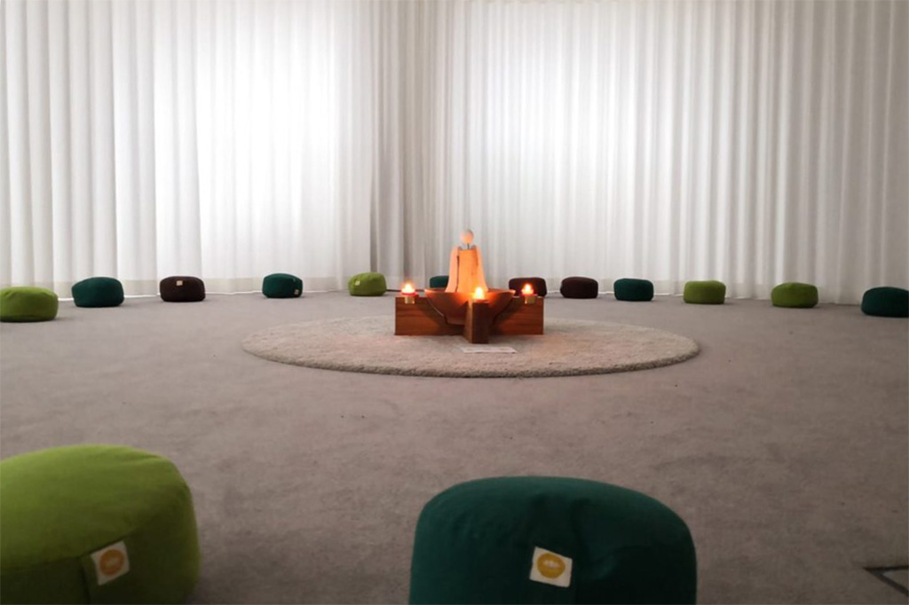 Raum der Stille - Grüne Kissen liegen im Kreis auf einem Teppichboden, in der Mitte stehen ein Holzengel und vier brennende Kerzen